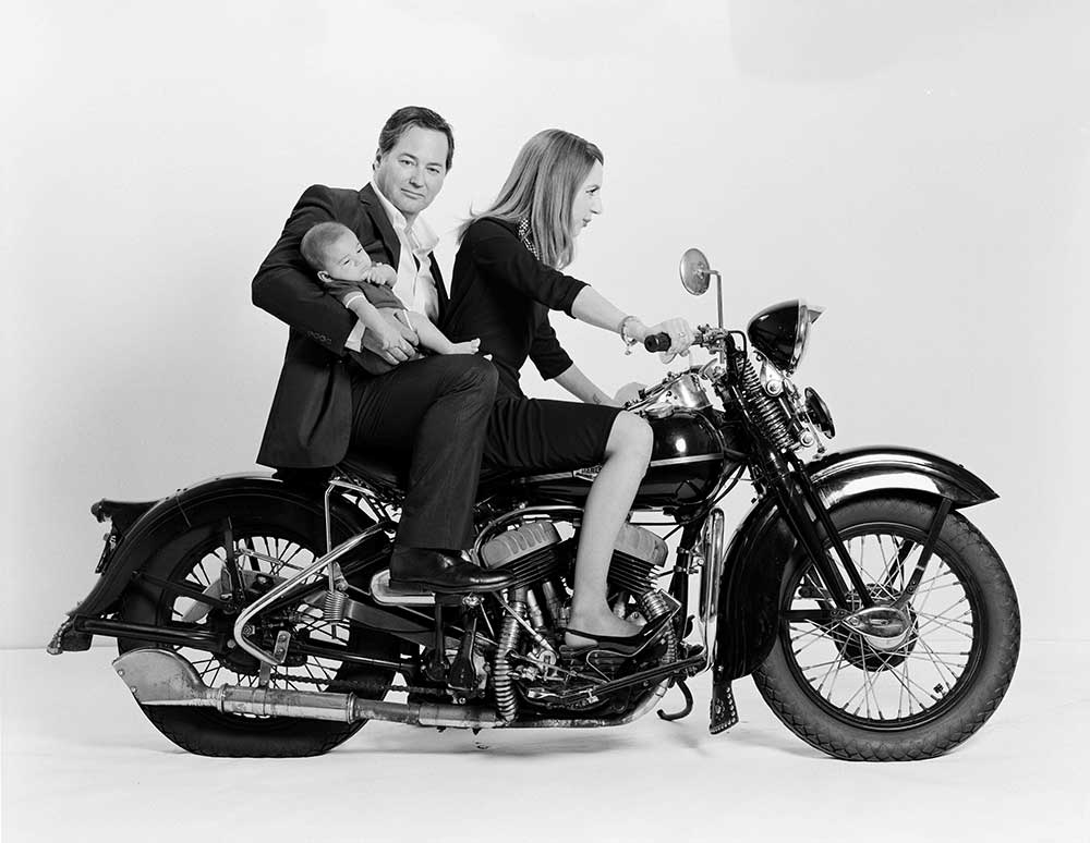 Black-and-white photo of Blair Walker in a suit posing on the back of a motorcycle with his partner in the front and infant in his arms.