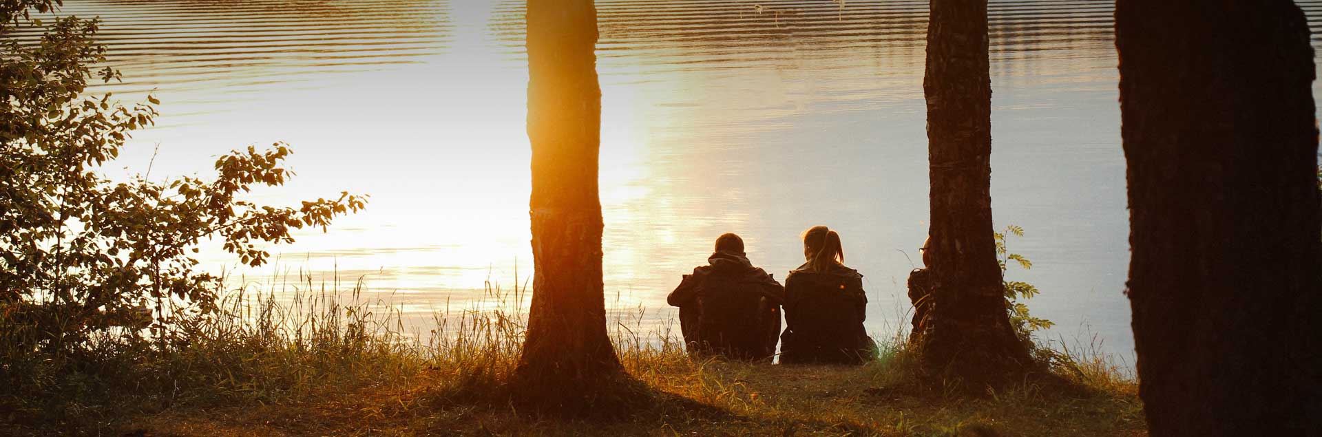 A man and woman sit side-by-side at the shoreline of a lake during a sunset.